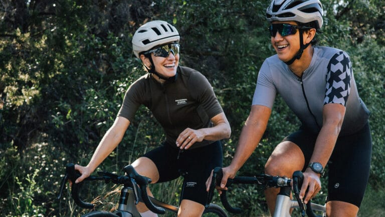 Two cyclists riding side by side on a forest road, smiling and wearing helmets and sunglasses, representing TrainingPeaks coaching and endurance training experience